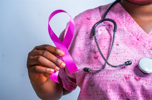 A care provider holds up a pink ribbon symbolizing breast cancer awareness and wears a stethoscope over pink patterned scrubs featuring hearts, flowers and a collage of positive words like joy, peace, happiness and care.